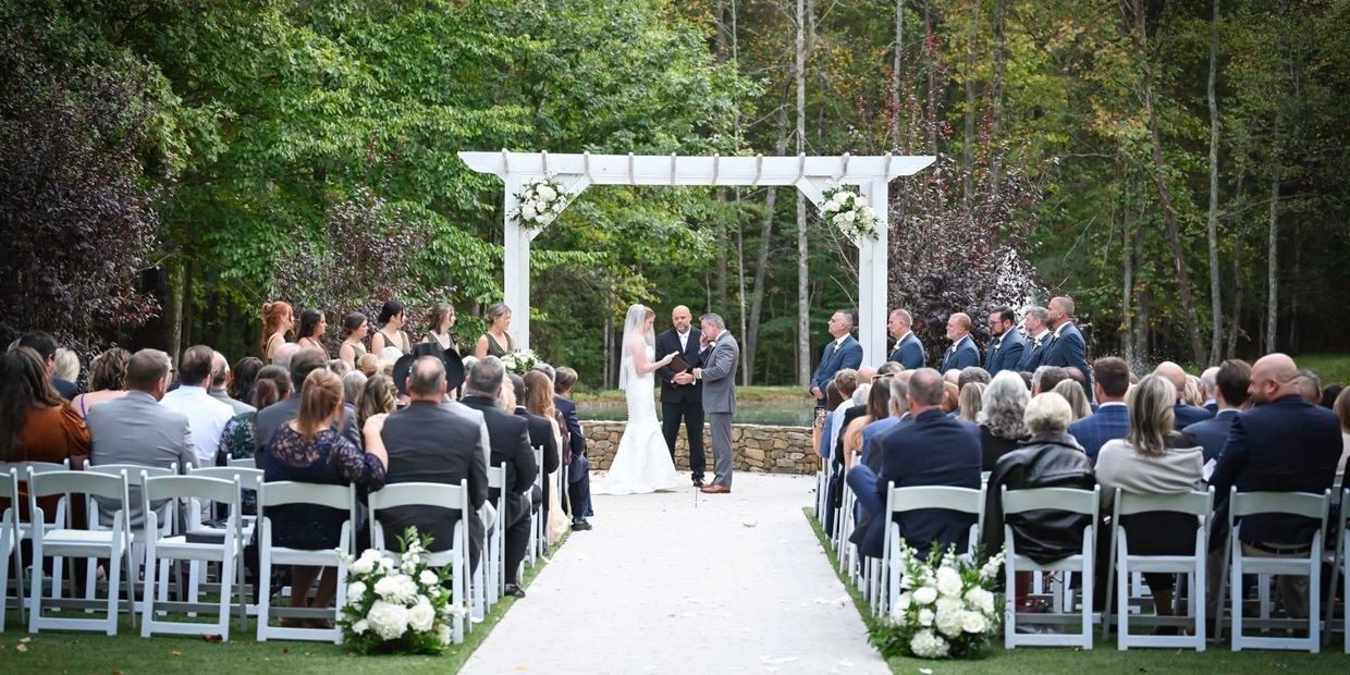 Outdoor wedding ceremony with bride, groom, officiant, and guests seated facing a white floral arch.