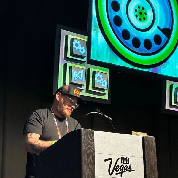 A man speaking at a Las Vegas conference podium with tech-themed visuals.