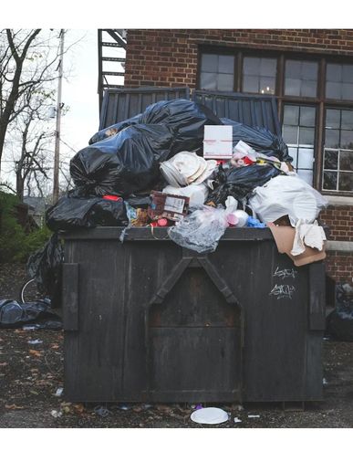 Overflowing dumpster filled with black garbage bags and trash items outside a brick building.