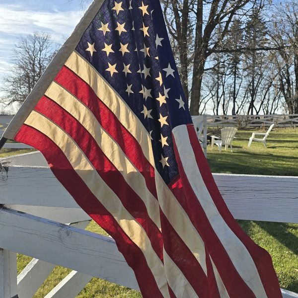 A worn American flag hanging on a white wooden fence in a sunny backyard.