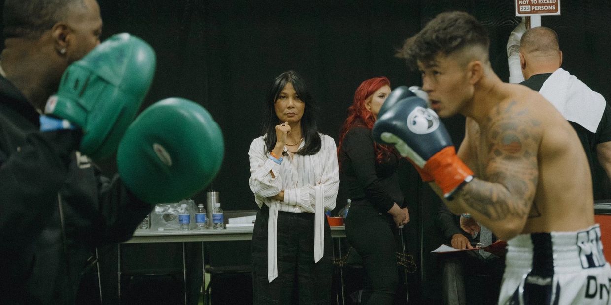 A boxer trains intensely with a coach while spectators watch in the background.