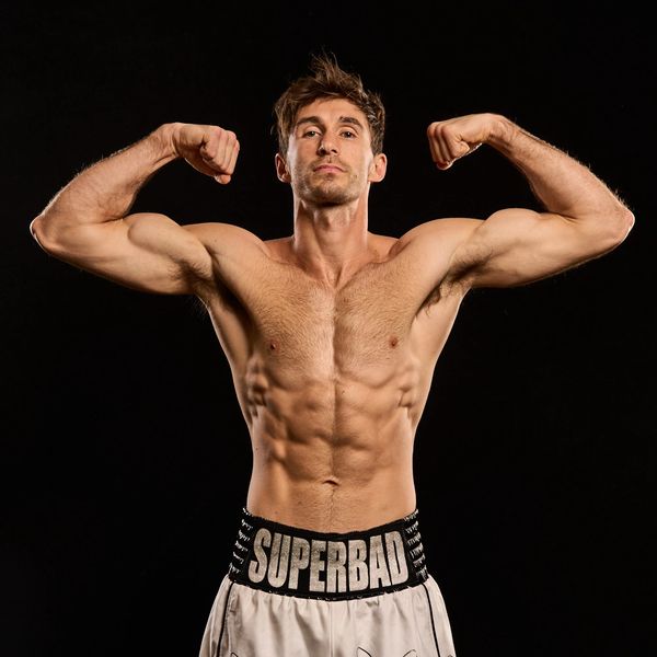Muscular man flexing biceps wearing 'SUPERBAD' shorts against black background.