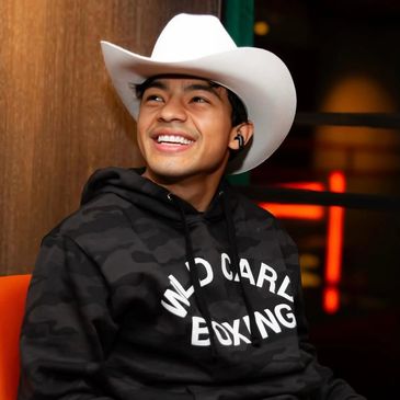A young man smiles wearing a white cowboy hat and a black hoodie.
