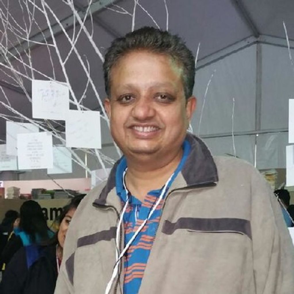 Smiling man in a jacket at an indoor event with hanging notes in the background.