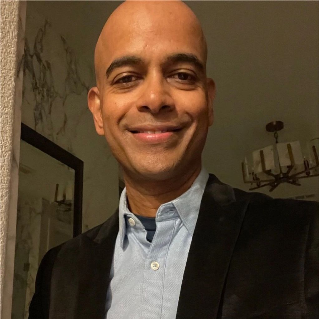 Smiling man wearing a light blue shirt and black jacket indoors.
