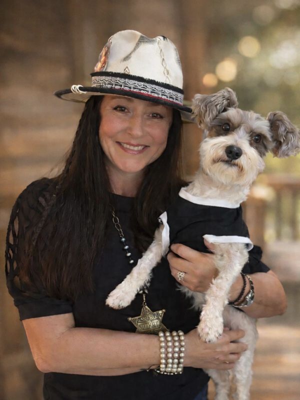 Woman in a hat holding a small dog dressed in black.