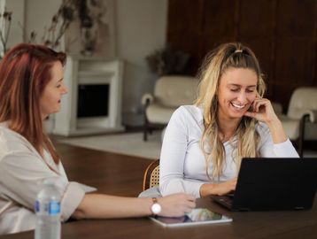 Two women smiling and working together at a laptop in a cozy room.