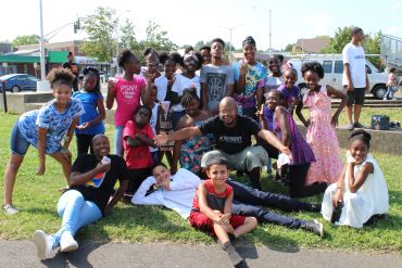 A diverse group of smiling children and adults posing outdoors on a sunny day.