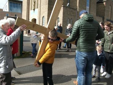 A boy carries a wooden cross outdoors with people around.