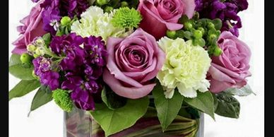 A vibrant bouquet of purple roses, white carnations, and green accents in a glass vase.
