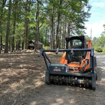 Orange skid steer with attachment clearing land in forested area.
