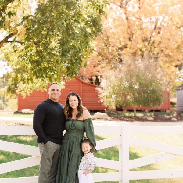 Family posing outdoors by a white fence on a sunny autumn day.