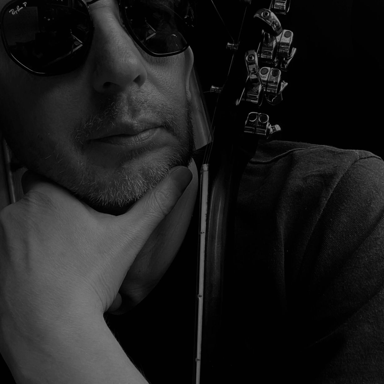 Man with sunglasses resting chin on hand next to a guitar headstock in black and white.