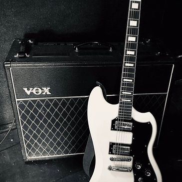 White electric guitar leaning against a VOX amplifier in a dimly lit room.