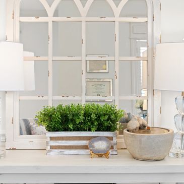 Decorative green plant and pottery on a white table in front of a large arched window mirror.