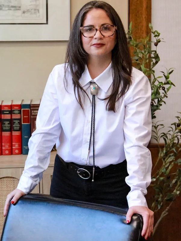 Woman in white shirt and bolo tie standing behind chair.