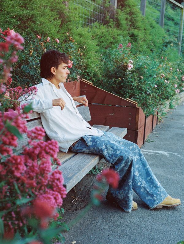 A person in a white shirt and patterned pants relaxes on a bench surrounded by flowers.