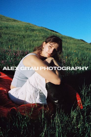 Young woman sitting outdoors on a red blanket in a grassy field under clear blue sky.