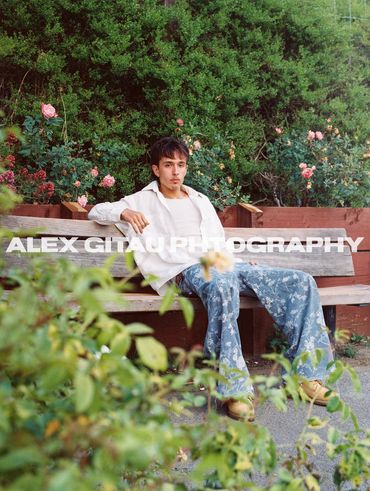 Young man sitting on a bench surrounded by lush greenery and flowering plants.