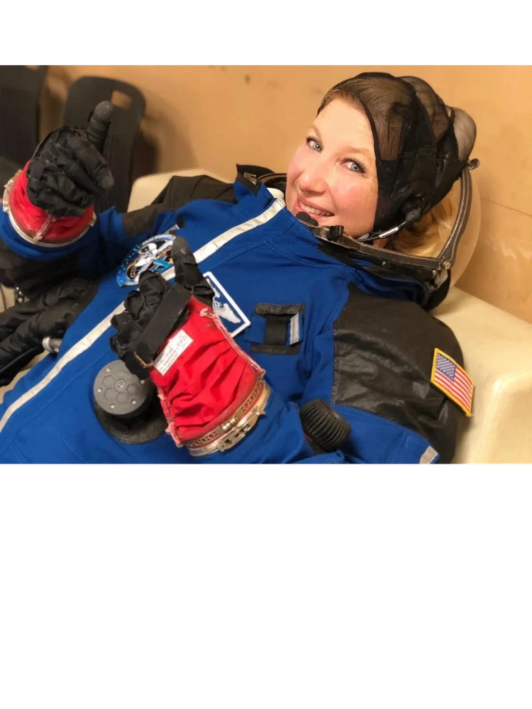 Kim Ellis Hayes Smiling astronaut in blue spacesuit giving thumbs up while reclining.