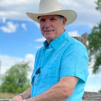 Older man in blue shirt and cowboy hat smiling outdoors.