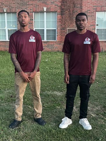 Two young men posing outdoors in maroon shirts and casual pants.