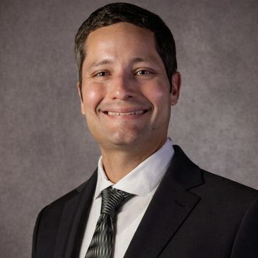 Man in a black suit and tie smiling against a gray backdrop.