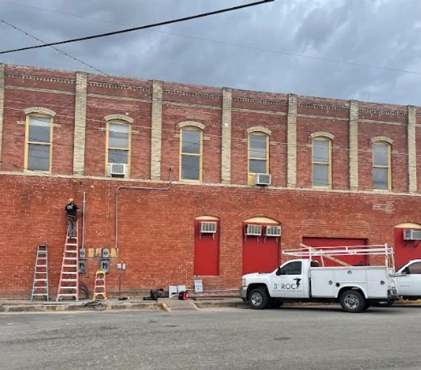Man on a ladder working on a red brick building with trucks parked nearby under cloudy sky.