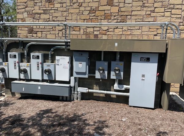 A row of electrical panels and meters mounted on a stone wall outside.