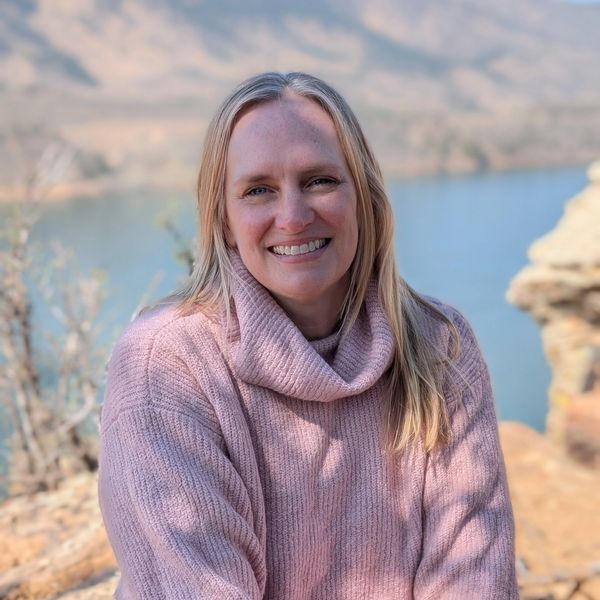 A woman in a pink sweater smiles outdoors near a body of water.