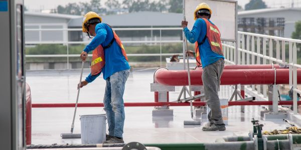 Two construction workers applying coating on a rooftop, wearing safety gear.