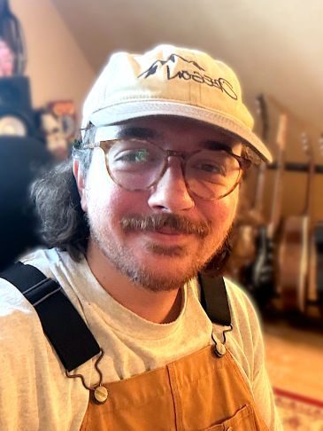 Man smiling with glasses, hat, and brown overalls.