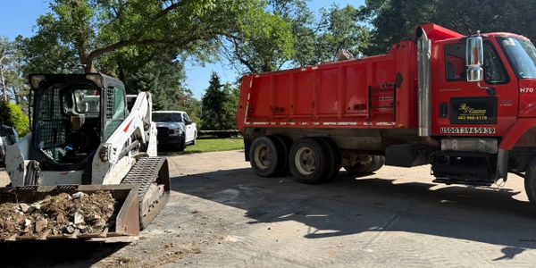 A white skid steer loader and a red dump truck on a sunny driveway surrounded by trees.
