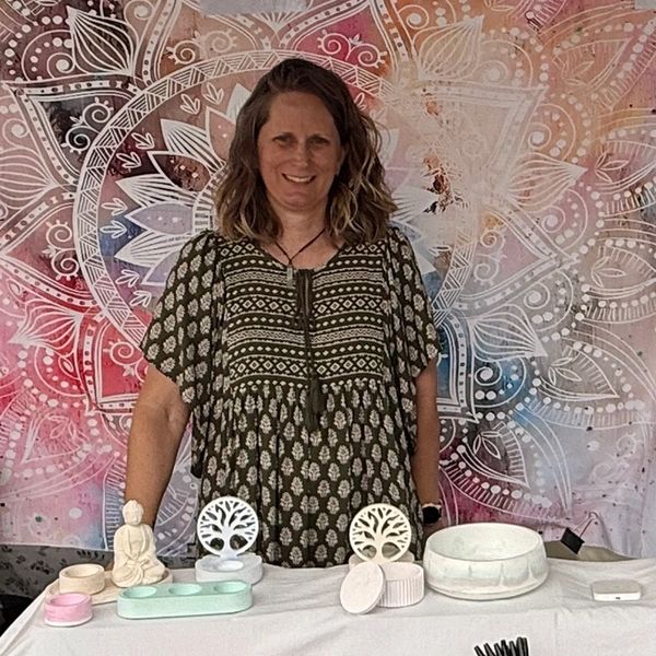 Woman standing behind a table with spiritual and wellness items against a colorful mandala backdrop.
