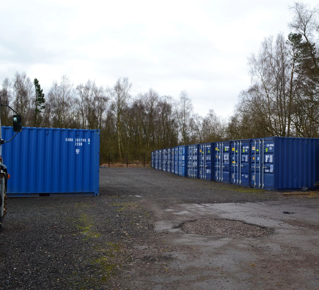 Blue shipping containers lined up on a gravel lot with trees in the background.