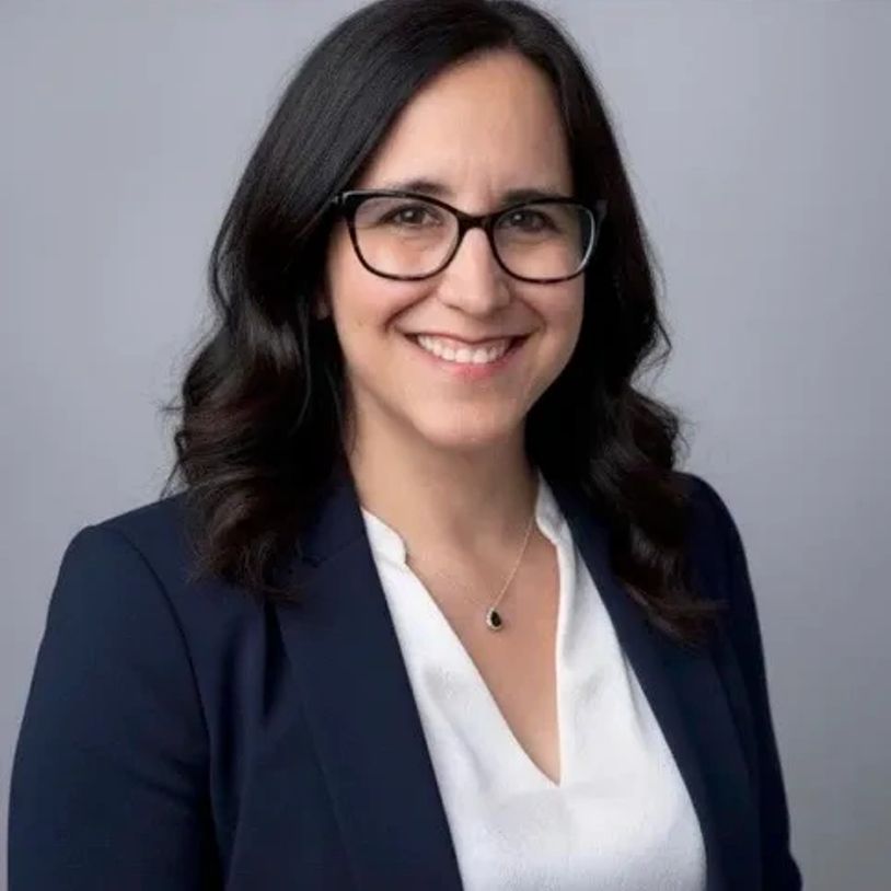 Professional woman with glasses smiling in a blazer and white blouse.