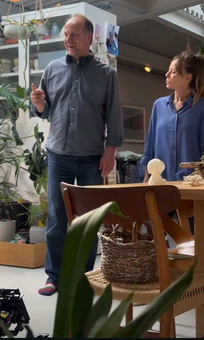 Two people talking indoors near plants and furniture.