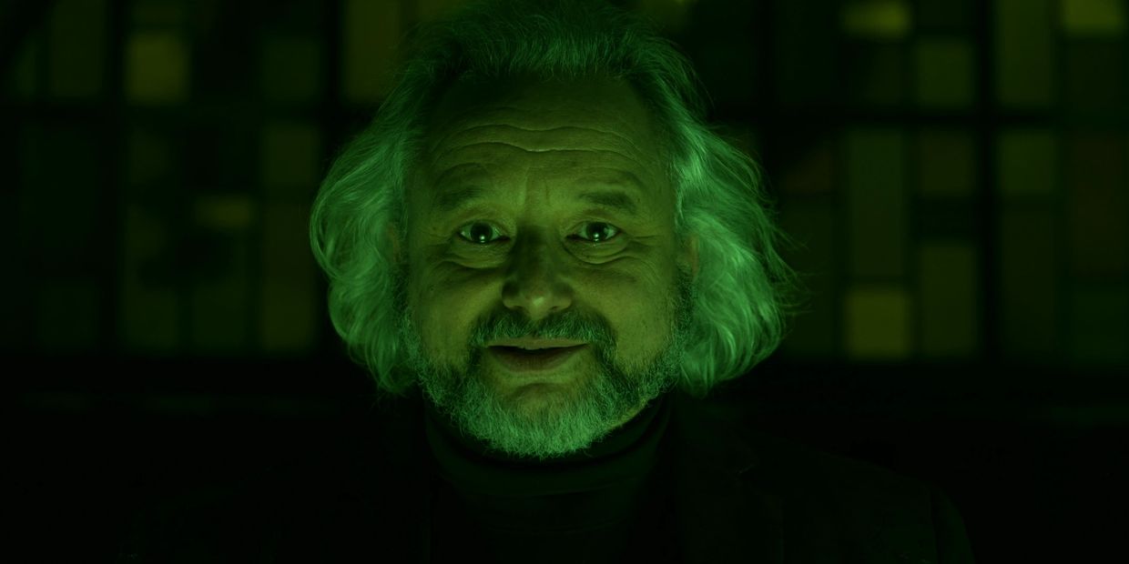 Older man with white hair and beard, lit by eerie green light, smiling slightly.