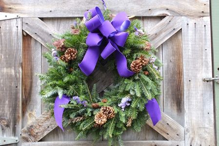 Green wreath with pinecones and purple ribbon on wooden door.