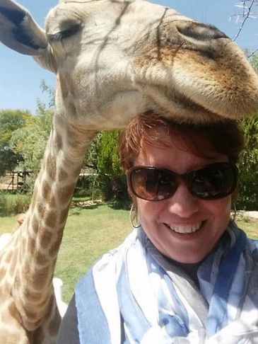 Woman wearing sunglasses takes a close selfie with a giraffe nuzzling her head.