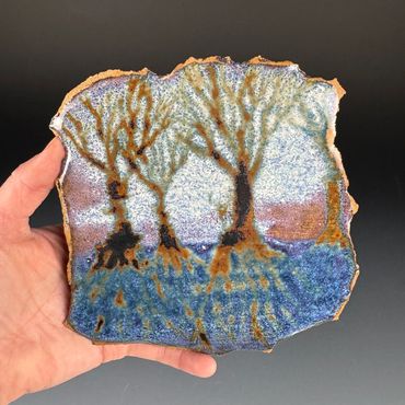 Hand holding a ceramic tile with a tree landscape design.