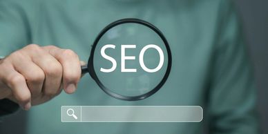 Person holding magnifying glass over the word SEO with a search bar below.