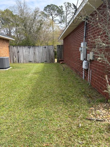 A backyard with green grass, a wooden fence, and a brick house wall with utility boxes.