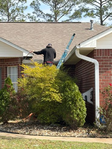 Man cleaning gutters on a sunny day with a ladder and hose.