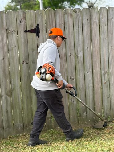 Person using a string trimmer to cut grass near a wooden fence.