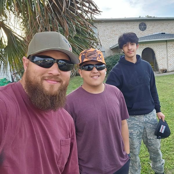 Three men smiling outdoors in casual wear with sunglasses and hats.