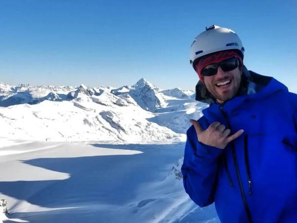 Smiling man in blue jacket and helmet poses on snowy mountain with clear blue sky.