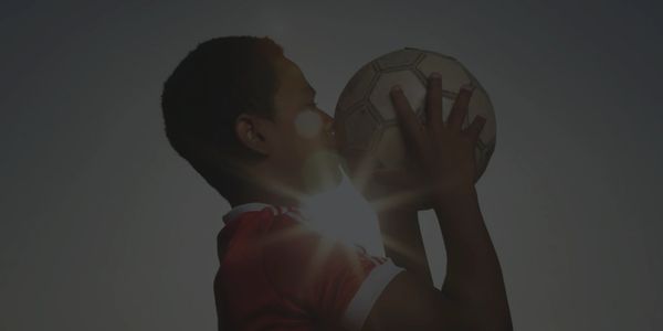 Boy in red jersey holding and kissing a soccer ball with sunlight behind.