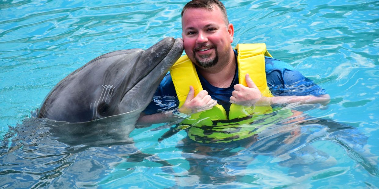 A man in a yellow life jacket smiling as a dolphin touches his face in a pool.