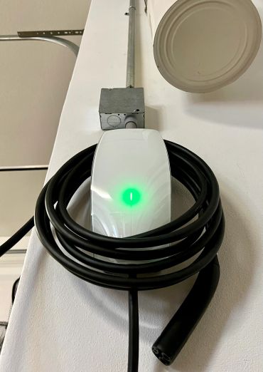 Wall-mounted electric vehicle charger with coiled black cable and glowing green indicator.
